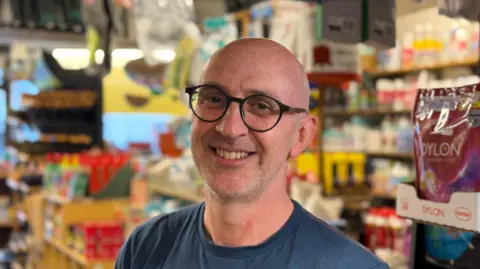Ben Hann looks at the camera. He wears glasses and a blue T-shirt. He stands in front of a wall of hardware goods in soft focus. 