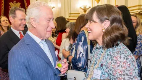 King Charles III on the left is wearing a blue suit with a thin white stripe, a white shirt and pale blue tie. He is holding a glass in which there is clear liquid and a slice of lime. He is looking at Caroline Jones and smiling with a closed mouth. Caroline on the right is smiling, she wears a light green floral dress with a long string of pearls and pearl earrings in a gold setting. 