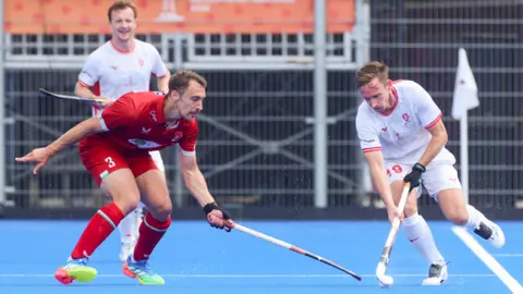 Getty Images A man wearing red on the left and another in white on the right are holding hockey sticks near the ground on a blue surface. Another man in white is in the background on the left.