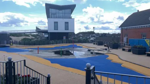 A largely empty paddling pool with a small amount of water in the bed. The floor is electric blue with a yellow border. In the background is a modern building dominated by a glass frontage.