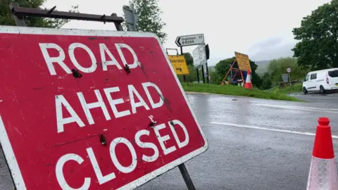 BBC/Seb Cheer Large red sign reading "road ahead closed" with a "diversion" sign in the background, next to a sign reading "Ingleton B6255".