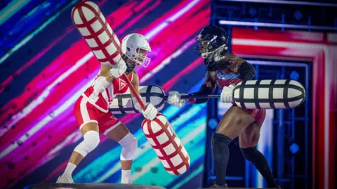 BBC/Hungry Bear Media Ltd/Graeme Hunter Two people holding giant sticks with foam ends, one person is wearing a red and white outfit, the other a red and blue outfit