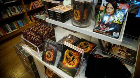 Getty Images A shelf in a bookshop loaded with Hunger Games books and stationery bearing the series logo of a golden bird inside a burning ring, and magazines bearing portraits of Jennifer Lawrence.
