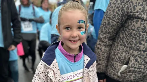 Family photogaph Elsie Dot Stancombe with blond hair in a pony tail and sparkly blue face sequins around her left eye. She is wearing a pale pink and navy coat with a blue running race top over a hot pink fleece. She is smiling. 