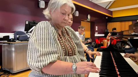 Alex Dunlop/BBC A woman is sitting at and playing a piano in a large room. She has short white hair and is wearing a white and brown patterned dress. Next to her, is a woman sitting watching. There are two people in the background playing instruments.