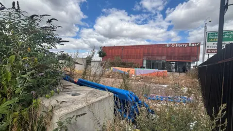 A large red metal structure in the background which has the sign Perry Barr is the railway station, in the foreground there are metal barriers on the right hand side and weeds and undergrowth on the concrete floor with lots of litter and detritus