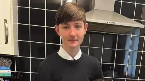 Jacob, 14, with brown hair wearing a white shirt and black crew neck jumper in a kitchen with black kitchen tiles on the wall behind him and an oven hob and kitchen cupboard.