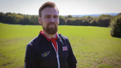 Karl Hinett, a man with swept-back reddish hair and a gingery beard, stands in a field. He is wearing a tracksuit top with a Union flag and and an Invictus Games badge.