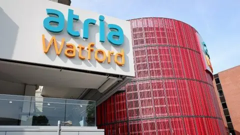 Getty Images The exterior of a modern shopping centre, with "atria Watford" in blue and orange lettering on a white panel above the glass wall of a shopping centre cafe. There is a circular red wall to the right.