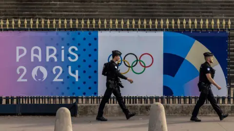 Getty Images Two armed police officers walk in front of a poster for the Paris Olympics. They are seen in a side profile. 