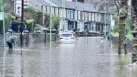 A flooded Berw Road in Pontypridd, with a car in water up to its wheels, a person up their knees, and a row of houses in the background