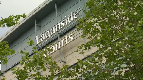 A silver sign for Laganside Courts on the side of a building partially obscured by tree branches