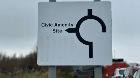 A large white road sign with a three-quarter circle indicating the civic amenity site is to the left. There is a queue of cars heading for the site on the right.