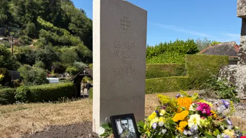 A headstone is pictured on a grave, besides a picture of a soldier and flowers
