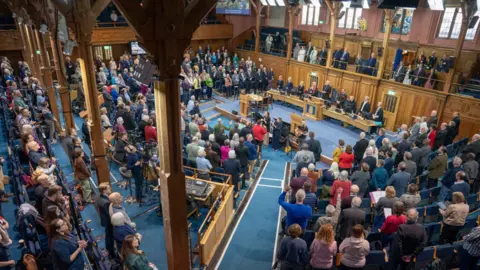 A large gathering is taking place inside a grand wood-paneled hall or assembly chamber. Hundreds of people are standing throughout the room, facing a central stage area where officials or speakers are seated behind long desks. The space has tall timber beams, balconies filled with attendees, stained-glass windows, and bright stage lighting. Near the centre floor, a small group of musicians appears to be performing, with microphones and instruments set up. Several audience members have their hands raised, suggesting a ceremonial, religious, civic, or commemorative event. Camera operators and media equipment are visible along one side of the hall, indicating the event is being recorded or broadcast.