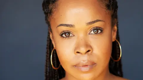 Claire Cousin Photography Azuka Oforka pictured her in a headshot. She is wearing gold hooped earrings and her hair is in braids. 