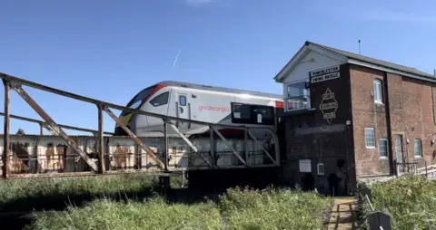 Andrew Turner/BBC A Greater Anglia train crosses a swing bridge. A brick building with white boards at the top is next to the crossing with the words Somerleyton Swing Bridge written on it. There is a path leading to the building with overgrown verges either side.