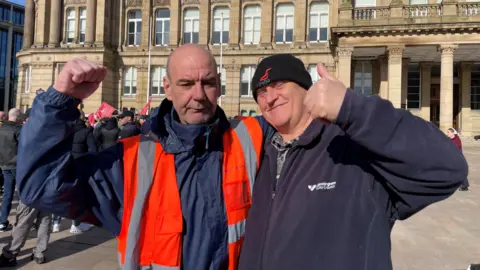 Dave and Johnny stood next to each other with an arm around each other's shoulders. Dave, wearing a high-vis orange jacket and blue coat, has a hand in the air. Johnny, on the right, has a hat on and is wearing a blue cardigan and is doing a thumbs-up sign with one hand. 