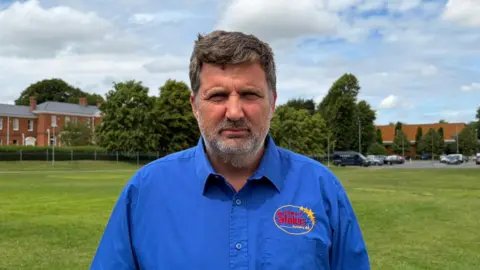 Edward Stokes Edward, wearing a blue shirt with his business logo on it. He has short cropped brown/grey hair and stubble, and is standing in a field