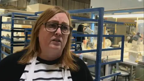 Sharon Yates has long red, brown hair just past her shoulders and wears black-rimmed glasses. She has a black top over a white and black striped top. Behind her are metal shelves, painted blue, to hold ceramics. Further back are rows of white mugs.