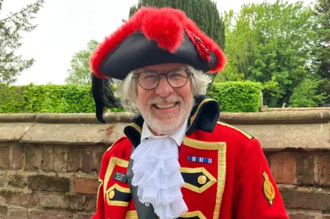 Henry Godfrey-Evans/BBC A man in a regal red town crier's outfit with a white plume coming from the neck. He has glasses and he's grinning