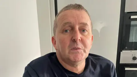 Mark Sneddon wearing a black t-shirt and looking at the camera. He has short light brown hair.