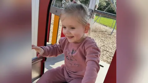 Sarah Normington A two-year old child with blonde hair is sitting on a small climbing frame in a play park. She is wearing a pink long-sleeved outfit with a rabbit on the front.