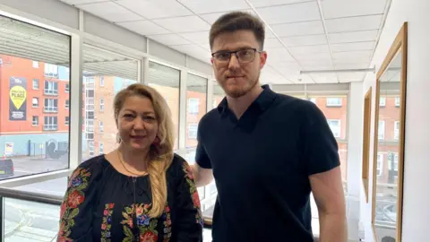 A woman has long blond hair over one shoulder, she is wearing large pink earrings and a black top with a traditional folk flower design. She is standing next to a man with short brown hair, a brown close shaved beard, glasses and a black top. They are standing on a mezzanine floor with large glass windows behind them