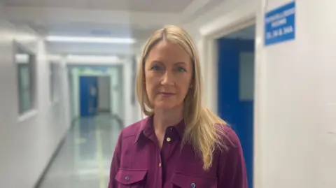 Sarah Parry, a woman with lond blonde hair and a maroon shirt, stands in a school corridor looking at the camera.