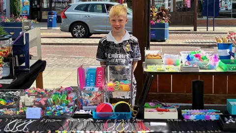 Blonde boy standing in front of stall with car behind on a town high street