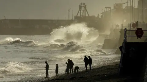beetlebug A group of silhouetted beach walkers are in the foreground with dramatic waves and sea spray behind them. 
