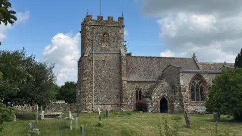 Seth Dellow Exterior of St Mary's church. A medieval stone church with a crenelated tower but no spire. The building sits within the grass churchyard where there are a number of upright and wonky gravestones.