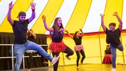 Keynsham Music Festival A group of young men and women do a Bollywood-style dance on a stage in a big yellow tent. The women wear black tops and leggings and grass skirts, while the men wear black T-shirts and jeans. 