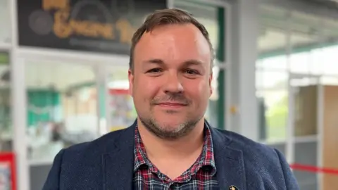 Shaun Whitmore/BBC Terry Jermy is standing looking towards the camera smiling. He has brown hair and eyes, and is wearing a blue and red chequered shirt, and a blue coat. He is standing in front off glass doors and windows. 