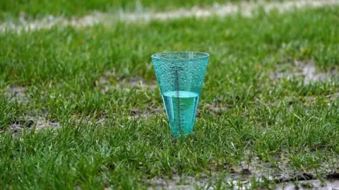 A clear plastic cup filling with rain on the side of a football pitch