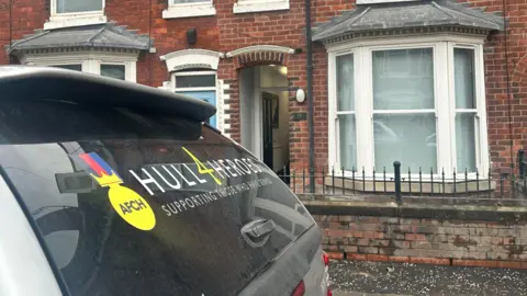 A close up of the back window of a car. On the rear window is a graphic reading: 'Hull4Heroes, supporting those who have served'. The car is parked in front of two terraced houses with bay windows.