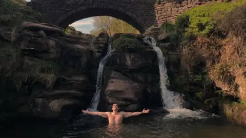 Nicola Byatt A topless man, his arms outstretched, in the water below a small waterfall