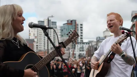 A man with red hair and a white T-shirt plays a guitar alongside a woman with blonde hair and a black top.