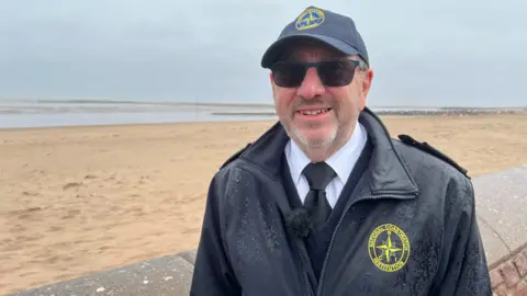 Malcolm is wearing dark shades and his navy blue unform with a branded waterproof coat. He is stood in front of Minehead beach.