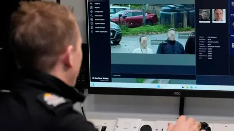 PA Media A police officer sitting at a computer terminal with his hand on a mouse. On the monitor in front of him is a video feed of live facial recognition, which shows two people walking past the camera with their faces pixellated. There is writing next to them which says "Unknown Face".