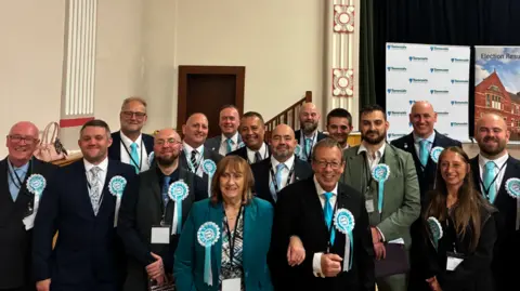 LDRS A group of people wearing suits and light blue ribbons stand smiling at the camera inside a hall. 