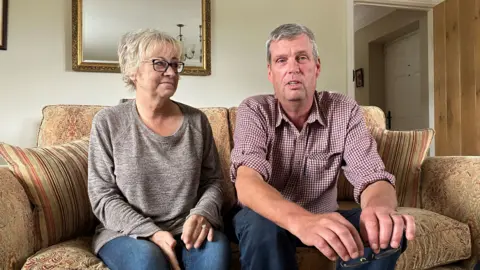 Amanda and Martin Beal are sitting on a brown sofa. They are a middle-aged couple and she is looking at him while he stares at the camera. 