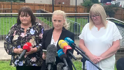 Three women, one with shoulder length brown hair and a fringe, another in a black suit with long blande hair tied up, and a third with long blonde hair with a fringe, standing beside each other in front of media microphones. 