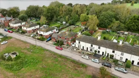 Dock Road in Sharpness, Gloucestershire. Terraced and semi-detached houses with gardens that reach trees can be seen lining one side of a street in a drone image. The River Severn can be seen in the background.