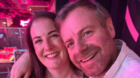 Hudgell Solicitors Nicholas Gibbs, in a room, with his arm around a woman. Both are smiling and the lights look pink. He has short fair hair and a beard and the woman long dark hair. 