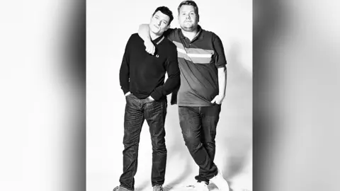 Tom Jackson James Corden and Matthew Horne in a black and white shot. Both are smiling at the camera standing with hands in their pockets. Gavin has his arm around Matthew's shoulder and neck.