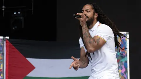 A man with long dreadlocks performs with a microphone while standing in front of a Palestine flag.