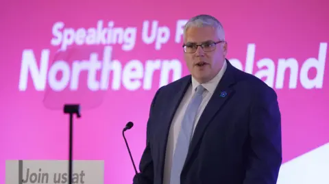 PA Media Gavin Robinson speaking at a microphone with a teleprompter in front of him. He is wearing a suit and tie. Behind him in large lettering it says "Speaking up for Northern Ireland"