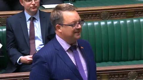 UK Parliament A man with short brown hair and glasses, wearing a navy pintstripe suit with lilac shirt and a purple tie. His body and face are angled away from the camera and he is speaking. In the background a man in a dark suit and maroon tie can be seen sitting on a row of green cushioned benches with ornate wooden carvings at the top.