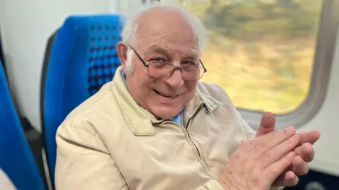 Tony is wearing a cream jacket and looking toward the camera smiling over the top of his glasses. He is clapping his hands in his train seat as the scenery behind blurs into the background. 